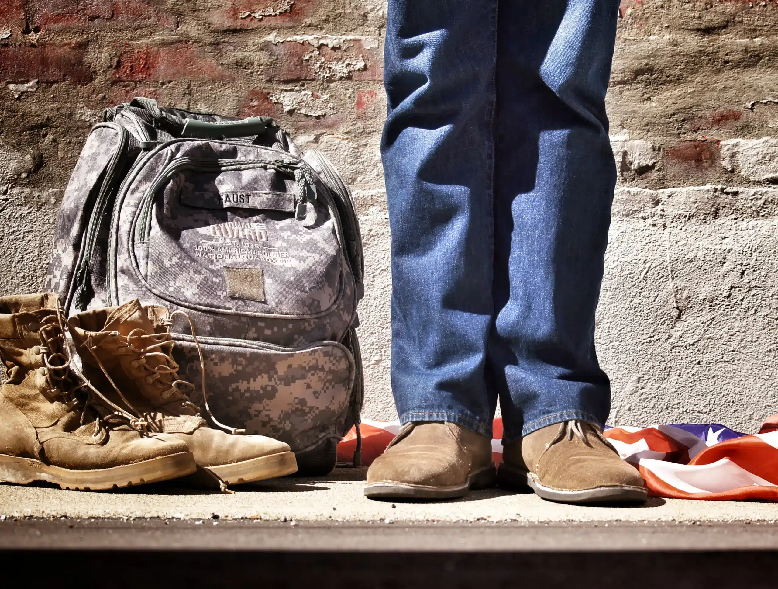 Man standing next to military backpack and boots with American flag at his feet behind him